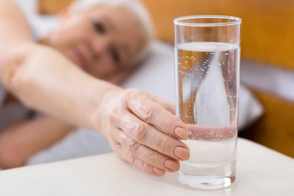 szklanka z wodą po którą sięga leżąca seniorka